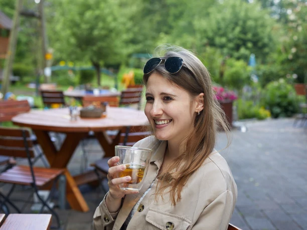 Junge Frau lächelt und hält ein Getränk im Freien, umgeben von Bäumen und Holzmöbeln.