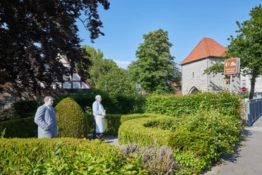 Zwei lebensgroße, weiß gestrichene Figuren vor einer historischen Kirche, umgeben von grüner Hecke.