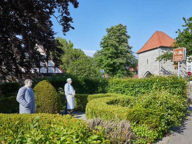 Zwei lebensgroße, weiß gestrichene Figuren vor einer historischen Kirche, umgeben von grüner Hecke.