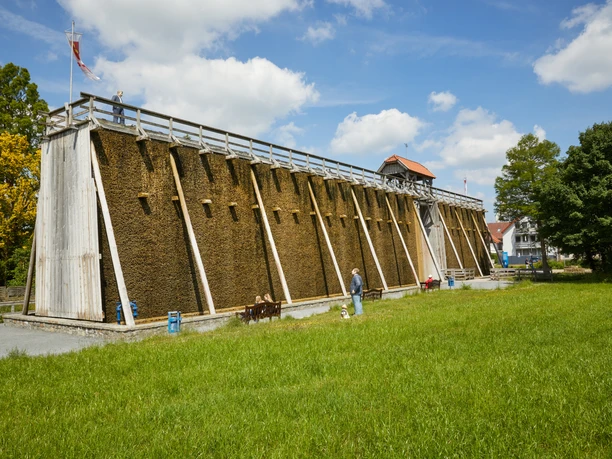 Ein Gradierwerk im Grünen; Besucher spazieren auf dem Weg daneben. Holzgerüst mit Reisig, klarer Himmel.