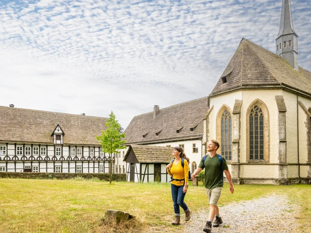 Lügde-Kloster Falkenhagen-Teutoburger-Wald-Tourismus-D-Ketz-098.jpg Zwei Wanderer vor einer historischen Kirche mit Fachwerkgebäuden unter blauem Himmel.