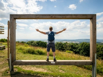 Mann mit Rucksack steht auf einer Holzplattform, ausgebreitet, mit Blick auf hügelige Landschaft.