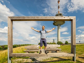 Ein Mann mit ausgebreiteten Armen steht auf einer Bank vor einem hölzernen Bilderrahmen im Freien. Im Hintergrund erhebt sich ein Fernsehturm unter einem blauen Himmel mit Wolken. Die Umgebung lädt zum Verweilen in der Natur ein.