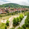 Luftaufnahme einer malerischen Stadt mit Fachwerkhäusern, Fluss und grüner Hügellandschaft im Hintergrund.