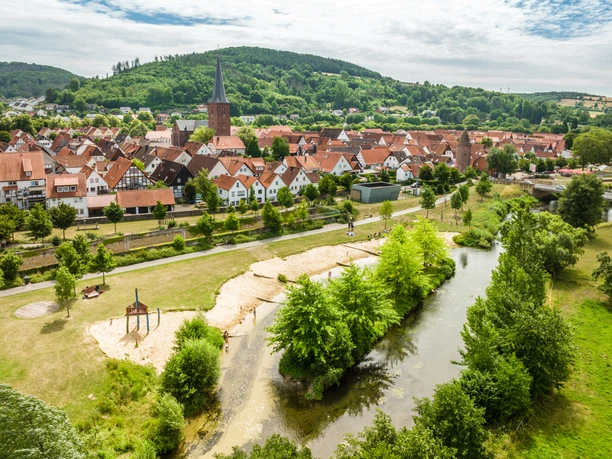 Luftaufnahme einer malerischen Stadt mit Fachwerkhäusern, Fluss und grüner Hügellandschaft im Hintergrund.