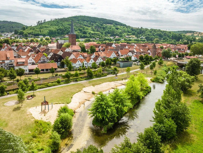 Lügde-Emmerauenpark-Teutoburger-Wald-Tourismus-D-Ketz-045.jpg Luftaufnahme einer malerischen Stadt mit Fachwerkhäusern, Fluss und grüner Hügellandschaft im Hintergrund.