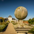 Eine historische Sonnenuhr aus Stein in einem gepflegten Schlossgarten vor einem barocken Gebäude.