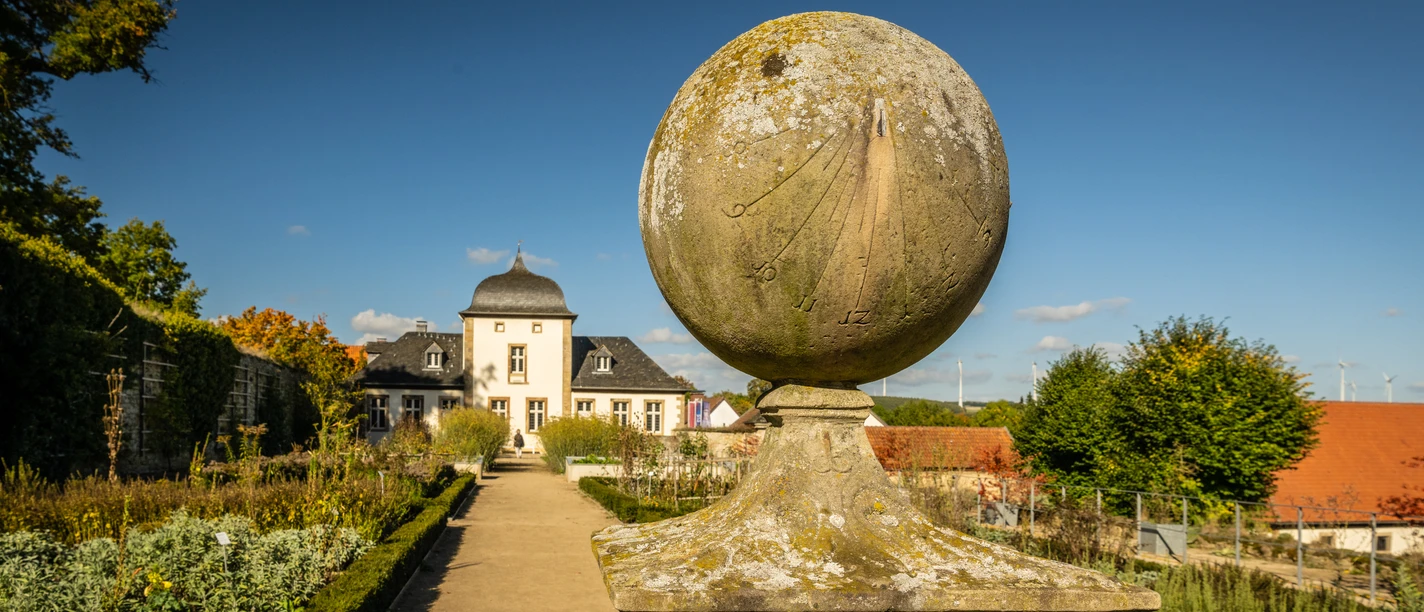 Eine historische Sonnenuhr aus Stein in einem gepflegten Schlossgarten vor einem barocken Gebäude.