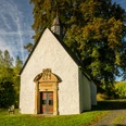 Eine kleine, historische Kapelle mit weißen Wänden, umgeben von grünen Bäumen und blühender Natur.
