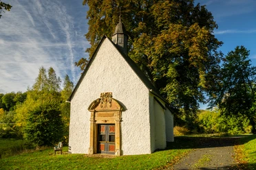 Lichtenau-Annenkapelle-Teutoburger-Wald-Tourismus-D-Ketz-007-CC-BY-SA.jpg Eine kleine, historische Kapelle mit weißen Wänden, umgeben von grünen Bäumen und blühender Natur.