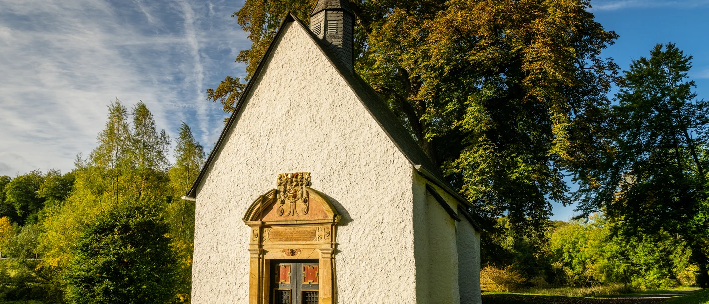 Lichtenau-Annenkapelle-Teutoburger-Wald-Tourismus-D-Ketz-007-CC-BY-SA.jpg Eine kleine, historische Kapelle mit weißen Wänden, umgeben von grünen Bäumen und blühender Natur.