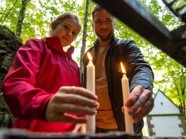Ein Paar beim Anzünden von Kerzen in einem Wald, im Hintergrund eine kleine Kapelle.