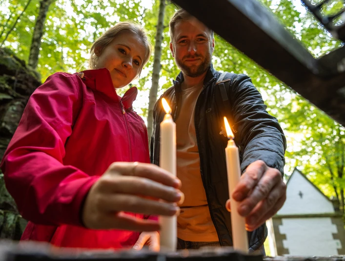 Ein Paar beim Anzünden von Kerzen in einem Wald, im Hintergrund eine kleine Kapelle.