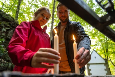 Ein Paar beim Anzünden von Kerzen in einem Wald, im Hintergrund eine kleine Kapelle.