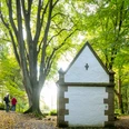 Kleine weiße Kapelle im Wald, große Bäume drumherum; Spaziergänger auf einem Laub bedeckten Weg.
