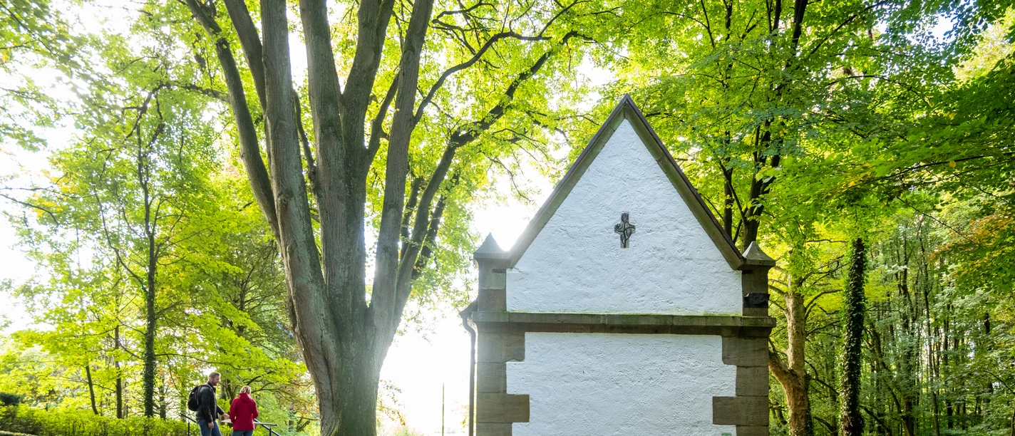 Kleine weiße Kapelle im Wald, große Bäume drumherum; Spaziergänger auf einem Laub bedeckten Weg.