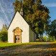 Zwei Radfahrer passieren eine kleine, weiße Kapelle in einer herbstlichen Landschaft bei sonnigem Wetter.