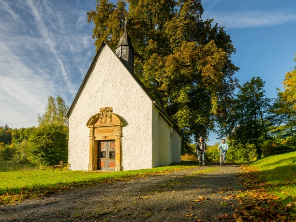 Lichtenau-Annenkapelle-Teutoburger-Wald-Tourismus-D-Ketz-009.jpg Zwei Radfahrer passieren eine kleine, weiße Kapelle in einer herbstlichen Landschaft bei sonnigem Wetter.