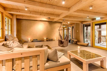 Cozy room with wooden furniture, hanging chair, candles and cushions on a sofa; warm lighting and large windows with a view outside.