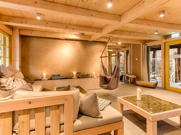 Cozy room with wooden furniture, hanging chair, candles and cushions on a sofa; warm lighting and large windows with a view outside.