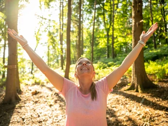Kalletal-Waldbadenpfad Heidelbeck-Teutoburger-Wald-Tourismus-D-Ketz-133.jpg Eine Frau steht im Wald, lächelt und hebt beide Arme in die Luft, Sonnenlicht fällt durch die Bäume.