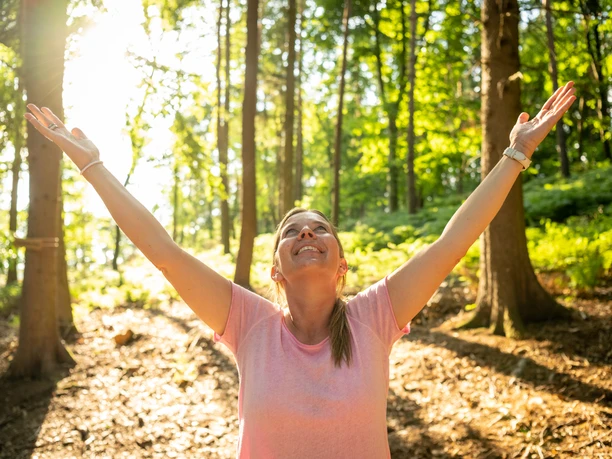 Kalletal-Waldbadenpfad Heidelbeck-Teutoburger-Wald-Tourismus-D-Ketz-133.jpg Eine Frau steht im Wald, lächelt und hebt beide Arme in die Luft, Sonnenlicht fällt durch die Bäume.