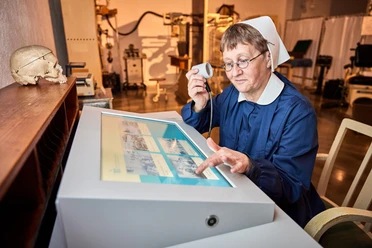 Medizinhistorische Sammlungen Eine ältere Frau in blauer Uniform und Haube bedient einen interaktiven Bildschirm und hält ein Mikrofon; ein Schädel liegt auf einem Regal daneben.