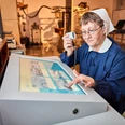Medizinhistorische Sammlungen Eine ältere Frau in blauer Uniform und Haube bedient einen interaktiven Bildschirm und hält ein Mikrofon; ein Schädel liegt auf einem Regal daneben.