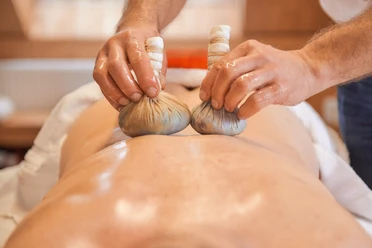 Eén persoon krijgt een massage met twee kruidenstempels op de rug; de sfeer is ontspannend en rustgevend.