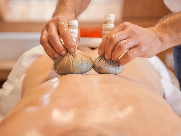 Eén persoon krijgt een massage met twee kruidenstempels op de rug; de sfeer is ontspannend en rustgevend.