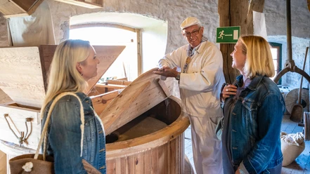 Porta Westfalica-Windmühle Holzhausen-Teutoburger-Wald-Tourismus-D-Ketz-027.jpg Drei Personen in traditioneller Kleidung stehen in einer historischen Mühle, umgeben von Holzelementen.