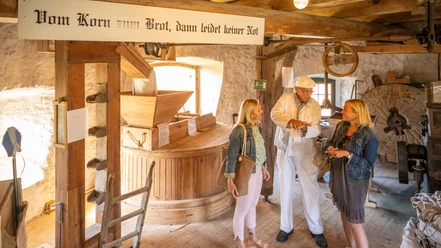 Eine historische Getreidemühle, in der ein Müller zwei Besucherinnen die Kunst des Mahlens erklärt.
