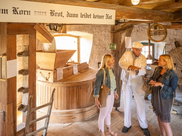 Eine historische Getreidemühle, in der ein Müller zwei Besucherinnen die Kunst des Mahlens erklärt.