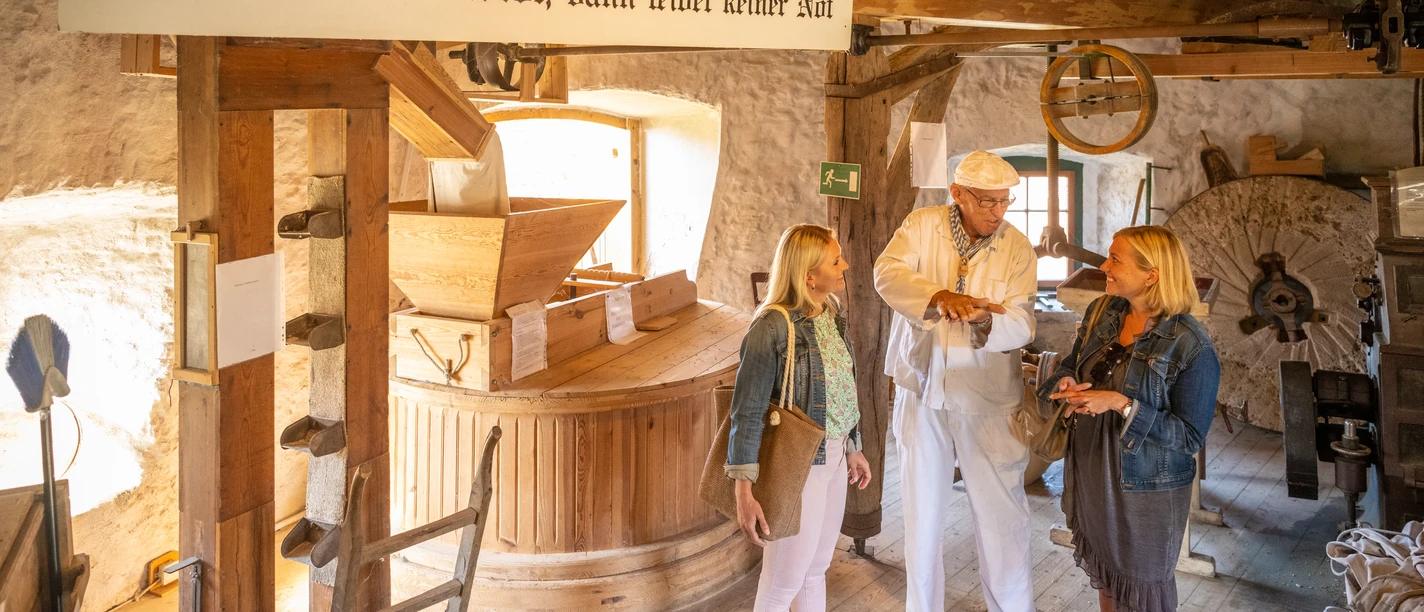 Porta Westfalica-Windmühle Holzhausen-Teutoburger-Wald-Tourismus-D-Ketz-032.jpg Eine historische Getreidemühle, in der ein Müller zwei Besucherinnen die Kunst des Mahlens erklärt.