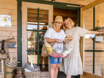 Minden-Biohof Kinkelbur-Teutoburger-Wald-Tourismus-D-Ketz-078.jpg Zwei Frauen stehen lachend vor einem Laden mit Holzfassade, eine hält einen Einkaufskorb mit Waren.