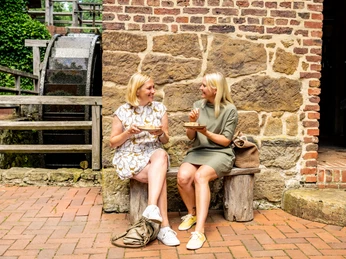 Bad-Oeynhausen-Wassermühle Bergkirchen Holzhausen-Teutoburger-Wald-Tourismus-D-Ketz-060.jpg Zwei Frauen in Sommerkleidern sitzen auf einer Bank vor der historischen Wassermühle Bergkirchen in Bad Oeynhausen