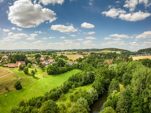 Bad-Oeynhausen-Wassermühle Bergkirchen Holzhausen-Teutoburger-Wald-Tourismus-D-Ketz-064.jpg Ländliche Landschaft mit Feldern, einem Fluss und Siedlung, umgeben von hügeligen Wäldern unter wolkigem Himmel.