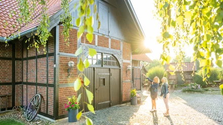 Minden-Windmühle Dützen-Teutoburger-Wald-Tourismus-D-Ketz-005.jpg Zwei Frauen stehen vor einem charmanten Fachwerkhaus im Sonnenschein, umgeben von grünen Blättern.