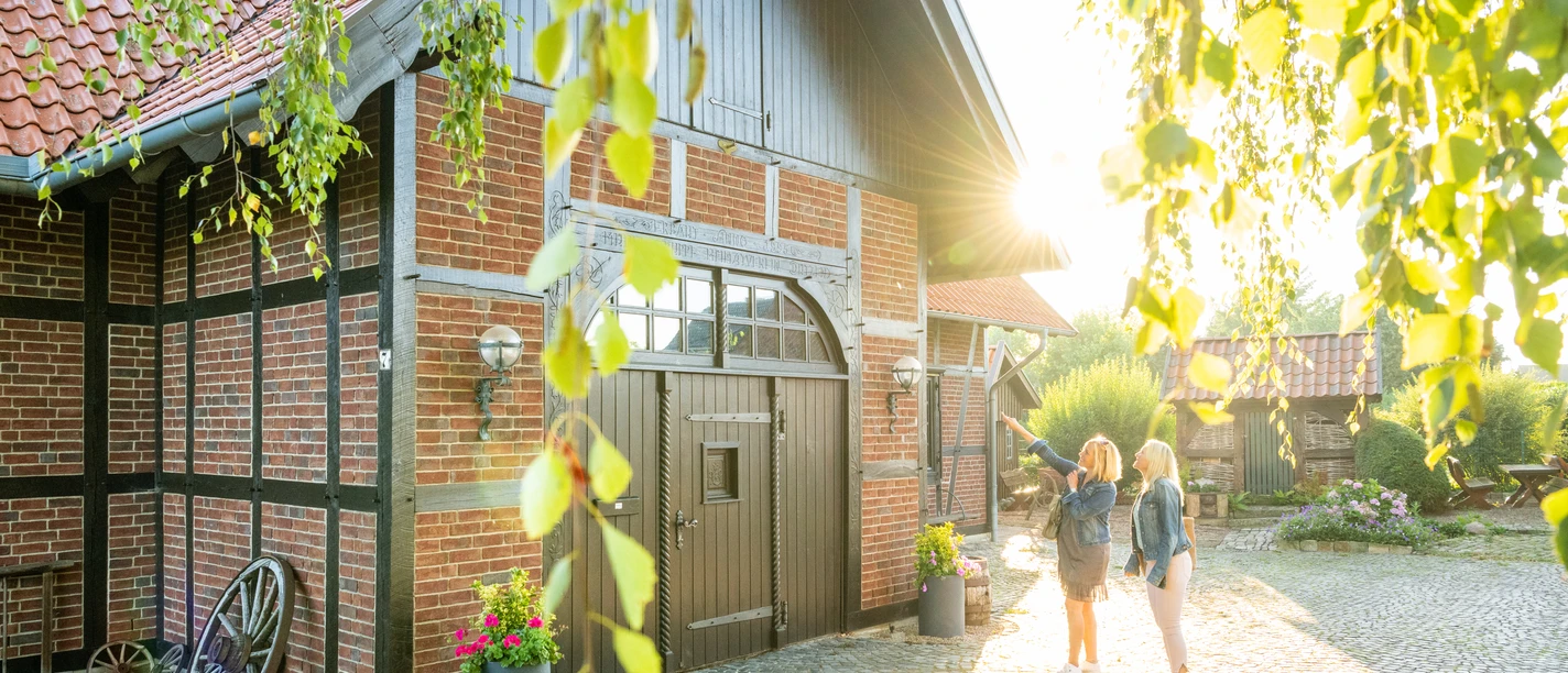Minden-Windmühle Dützen-Teutoburger-Wald-Tourismus-D-Ketz-005.jpg Zwei Frauen stehen vor einem charmanten Fachwerkhaus im Sonnenschein, umgeben von grünen Blättern.