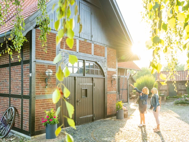 Minden-Windmühle Dützen-Teutoburger-Wald-Tourismus-D-Ketz-005.jpg Zwei Frauen stehen vor einem charmanten Fachwerkhaus im Sonnenschein, umgeben von grünen Blättern.