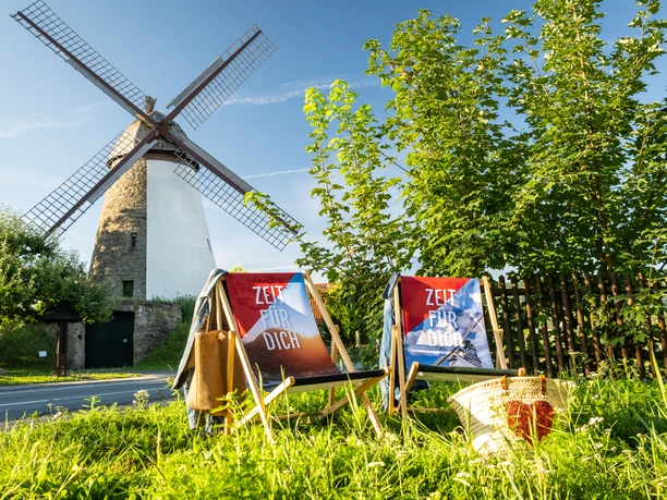 Zwei Liegestühle auf grünem Gras vor einer historischen Windmühle mit blauen Himmel im Hintergrund.