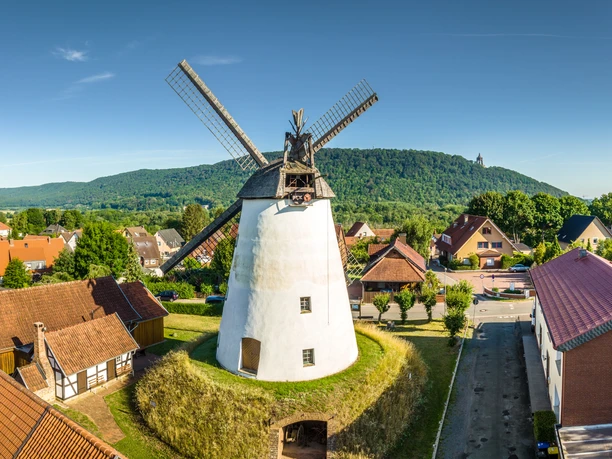 Weißer Turmholländer mit großen Flügeln inmitten eines Dorfes, umgeben von Bäumen und Hügeln.