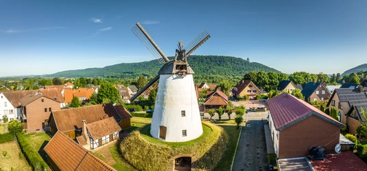 Porta Westfalica-Windmühle Holzhausen-Teutoburger-Wald-Tourismus-D-Ketz-022.jpg Weißer Turmholländer mit großen Flügeln inmitten eines Dorfes, umgeben von Bäumen und Hügeln.