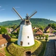 Porta Westfalica-Windmühle Holzhausen-Teutoburger-Wald-Tourismus-D-Ketz-022.jpg Weißer Turmholländer mit großen Flügeln inmitten eines Dorfes, umgeben von Bäumen und Hügeln.