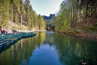 Amselsee Kurort Rathen A tranquil lake with green pedal boats on the shore, surrounded by dense forests and a clear sky, creates a peaceful atmosphere.