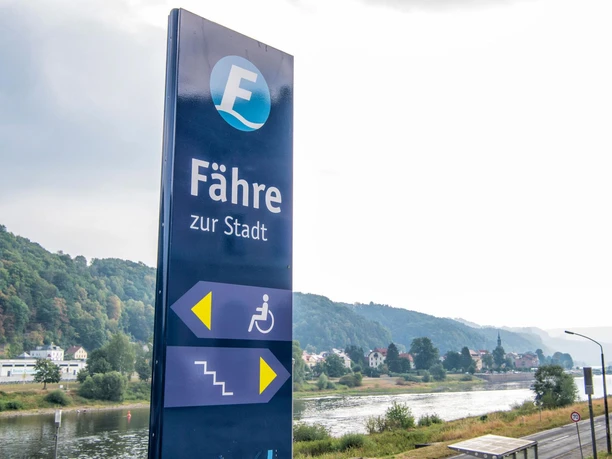 Fähre Bad Schandau Blaues Schild mit der Aufschrift "Fähre zur Stadt" und Piktogrammen für Rollstuhlzugang und Treppen, im Hintergrund Fluss und bewaldete Hügel.