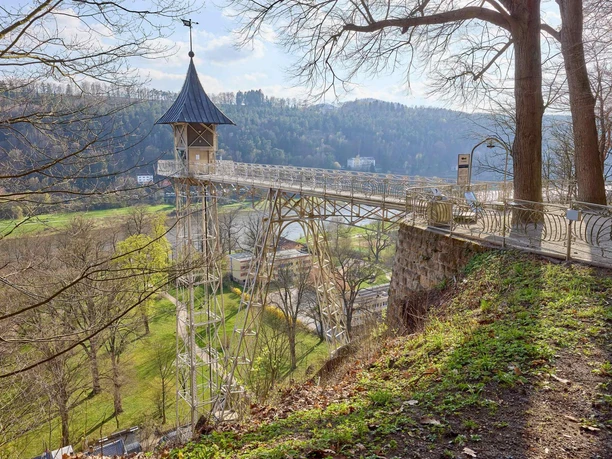 A metal bridge with a small tower leads over a green landscape with trees and hills in the background, in sunny weather.