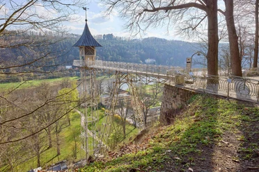 Historischer Personenaufzug Bad Schandau Eine Metallbrücke mit einem kleinen Turm führt über eine grüne Landschaft mit Bäumen und Hügeln im Hintergrund, bei sonnigem Wetter.