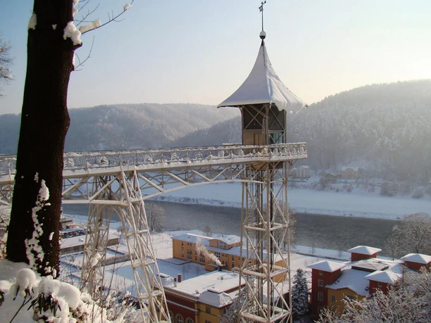 Historischer Personenaufzug Bad Schandau im Winter Ein schneebedeckter Aussichtsturm mit spitzem Dach steht über einem Fluss, umgeben von verschneiten Hügeln und bunten Gebäuden.