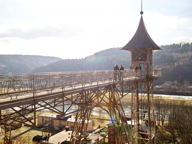 Historischer Personenaufzug in Bad Schandau Ein historischer Aussichtsturm mit spitzem Dach steht auf einer Brücke, darunter fließt ein Fluss; im Hintergrund bewaldete Hügel bei sonnigem Wetter.